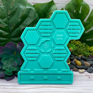 Puede incluir: Molde de silicona turquesa con forma de panal de abejas con un diseño de abeja en el centro. El molde tiene una base con tres secciones rectangulares. El molde está etiquetado como "DEFIANT ART CO."