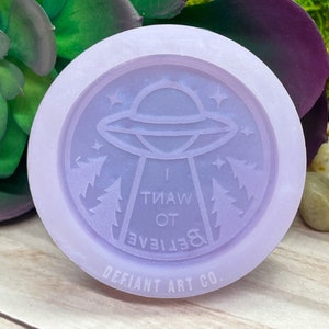 May include: Purple silicone mold with a raised design of a flying saucer, stars, and pine trees. The text "& Believe I Want To" is written in a circle around the design. The bottom of the mold reads "Defiant Art Co."