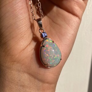 May include: A silver chain necklace with a teardrop-shaped opal pendant. The opal is iridescent with flashes of green, blue, and red. A small blue gemstone is set above the opal.