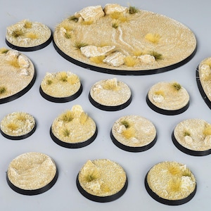 May include: A selection of miniature bases for tabletop games, showcasing a desert environment. The bases are in various shapes, with a tan surface, small rocks, and dry grass tufts. Each base has a black rim, suitable for miniature figures.