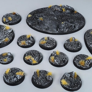 Puede incluir: Conjunto de bases de miniatura negras texturizadas, de diversas formas y tamaños. Cada base presenta mechones de color amarillo-marrón, creando un efecto de terreno. Las bases están diseñadas para figuras en miniatura, con formas redondas y ovaladas.