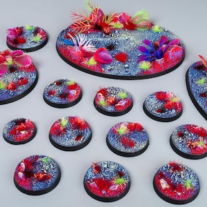 Puede incluir: Un conjunto de bases en miniatura para juegos de mesa. Las bases presentan un diseño vibrante y colorido con elementos rojos, azules y verdes, que se asemejan a un arrecife de coral submarino. Las bases varían en tamaño y forma, con un borde negro.
