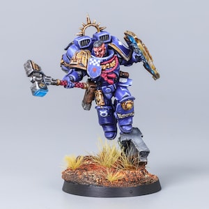 Peut inclure: Une figurine miniature peinte représentant un guerrier de l'espace en armure bleue avec des bordures dorées, tenant un grand marteau argenté à tête bleue lumineuse et un bouclier. La figurine a un détail de corde rouge et un socle texturé.