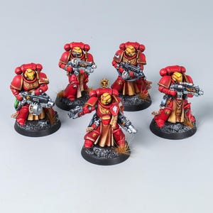 Puede incluir: Cinco figuras de soldados en miniatura con armadura roja y amarilla, cada una equipada con un arma de fuego futurista. Las figuras se alzan sobre bases negras circulares con detalles de suelo texturizados, acentos de armas en plata metálica y tabardos color canela.