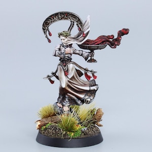 May include: A painted miniature figurine of a woman in a white robe holding a chalice with a red liquid pouring out. She is wearing a crown of leaves and has a crescent moon behind her head. The figurine is standing on a base with green grass and brown branches.