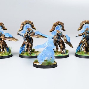 Könnte beinhalten: Miniaturfiguren von gepanzerten Figuren mit Gold- und Blauelementen, die auf kreisförmigen Sockeln mit Gras stehen. Eine Figur ist eine durchscheinende blaue Energieform. Die Figuren haben verzierte Kopfbedeckungen.