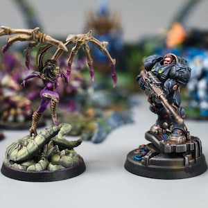 May include: Two painted miniature figures on circular bases. On the left, a purple-skinned creature with large, bony, tan wings stands on a green, textured base. On the right, a soldier in dark blue armor holds a rifle, standing on a metallic base.