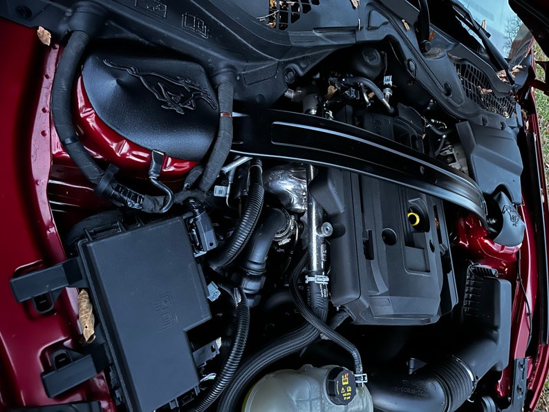 May include: Close-up view of the engine compartment of a red Ford Mustang. The engine is visible, along with various hoses and wires. The engine cover is black with a silver Ford Mustang logo.