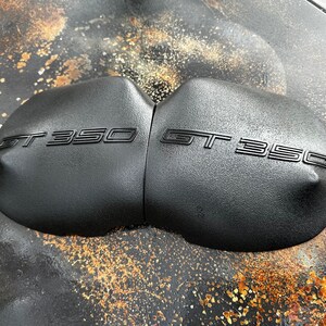 May include: Close-up of two black, rounded, textured motorcycle seat covers with the text "GT 350" embossed on each.