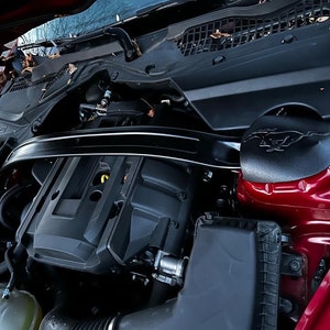 May include: Close-up view of a red car engine compartment. The engine is black and has a black plastic cover with a raised Ford Mustang logo. The engine is surrounded by various hoses and wires.