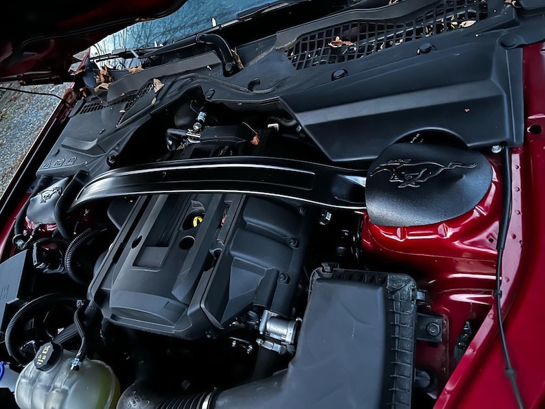 May include: Close-up view of a red car engine compartment. The engine is black and has a black plastic cover with a raised Ford Mustang logo. The engine is surrounded by various hoses and wires.