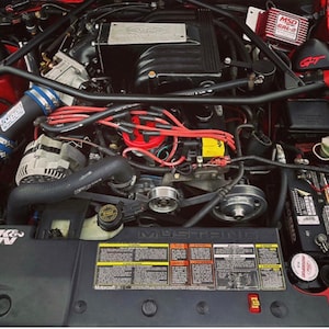 May include: A red car engine bay with a black engine, red and black wires, and a black air intake with a blue and silver filter. The engine has a black air filter with a K&N logo, a black battery with a red terminal, and a black and silver MSD 6AL-2 ignition box.