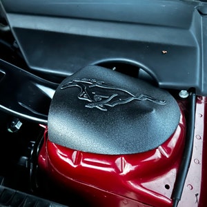 May include: Close-up of a black engine cover with a raised, silver Mustang logo. The cover is attached to a red car.
