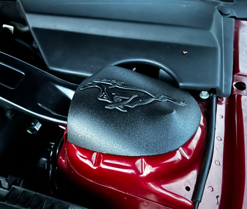 May include: Close-up of a black engine cover with a raised, silver Mustang logo. The cover is attached to a red car.