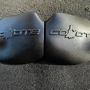 May include: Two black plastic engine covers with the word "Coyote" embossed on each. The word is in a stylized font with a coyote silhouette in the middle of the word.