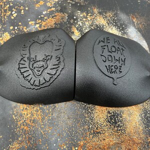 May include: Two black rubber car emblems with a 3D design. One emblem features a scary clown face with a red nose and a wide grin. The other emblem features a white balloon with the text "We All Float Down Here".