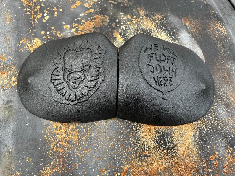 May include: Two black rubber car emblems with a 3D design. One emblem features a scary clown face with a red nose and a wide grin. The other emblem features a white balloon with the text "We All Float Down Here".
