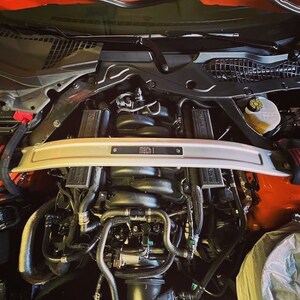 May include: Close-up view of the engine compartment of a red Ford Mustang Mach 1. The engine is visible, along with various hoses and wires. The car has a silver strut bar with the words "Mach 1" printed on it.