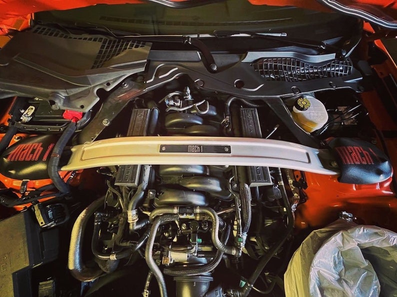 May include: Close-up view of the engine compartment of a red Ford Mustang Mach 1. The engine is visible, along with various hoses and wires. The car has a silver strut bar with the words "Mach 1" printed on it.