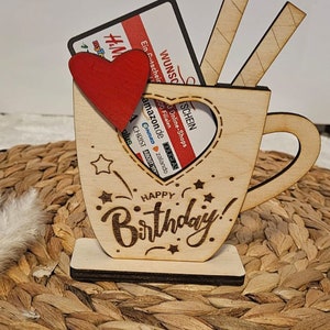 Könnte beinhalten: Holzbecher mit herzförmiger Öffnung, in der sich Geschenkgutscheine und ein rotes Herz befinden. Der Becher ist mit den Worten "Happy Birthday!" graviert und steht auf einer geflochtenen Oberfläche.