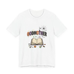 May include: A white short-sleeve t-shirt featuring a graphic with the text GODMOTHER in colourful letters above an open book. The design includes a spider web, a spider, bats, a black cat, a small pumpkin, a potion bottle, and a skull wand.