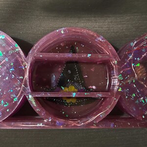 May include: A close-up of a three-sectioned, purple resin container with iridescent glitter. The center section holds a black and yellow design. The container is on a dark gray surface.