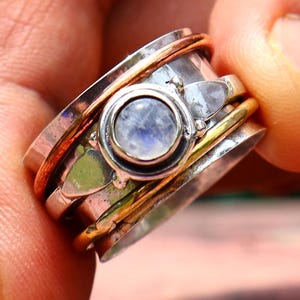 Pode incluir: Um anel giratório prateado e dourado com uma pedra preciosa central redonda azul clara. O anel tem várias faixas, incluindo detalhes em cobre e dourado. O anel é segurado contra um fundo verde e vermelho desfocado.