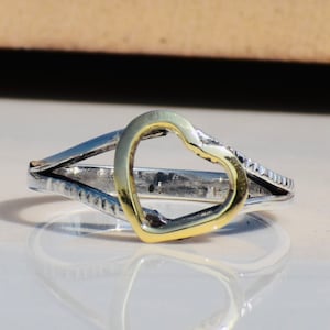 Op de afbeelding: Een zilveren ring met een gouden hartvormige uitsparing. De ring is getextureerd en heeft een delicaat ontwerp.