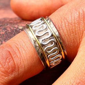 Peut inclure: Une bague tournante en argent et or avec un motif ondulé. La bague est portée au doigt d'une personne.