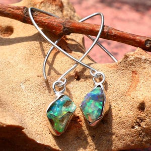 May include: Silver earrings featuring iridescent opal stones. The opals show green, blue, and yellow colours. The earrings are presented on a textured, tan surface with a brown twig.