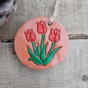 Puede incluir: Un adorno redondo de color terracota con un relieve de tres tulipanes rojos con tallos y hojas verdes. Se adjunta un lazo de cuerda natural para colgar. El adorno está sobre un fondo de madera rústica.