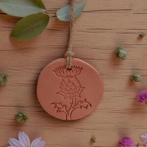 May include: A round, terracotta-colored ornament with a thistle design, suspended by a natural rope. The ornament is surrounded by flowers and greenery on a wooden surface.