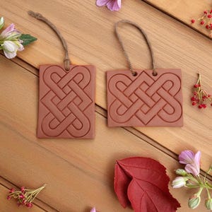 May include: Two rectangular, terracotta-colored ornaments with Celtic knot designs and twine loops for hanging. The ornaments are on a wooden surface, surrounded by flowers and red berries.