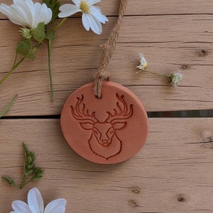 May include: A round, terracotta-colored ornament with a deer head design, hanging from a natural rope. White daisies and greenery are arranged around the ornament on a wooden surface, creating a rustic, nature-inspired aesthetic.