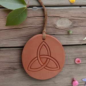 May include: A round, terracotta-colored pendant with a Celtic knot design, suspended by a natural twine cord. The pendant is set against a rustic wooden background, adorned with scattered flower petals and a sprig of leaves.