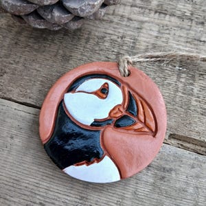 Puede incluir: Adorno redondo de color terracota con un relieve detallado de un frailecillo. El ave tiene la cara blanca, marcas negras y un pico naranja. Se adjunta una cuerda de yute para colgarlo. El adorno está sobre una superficie de madera.