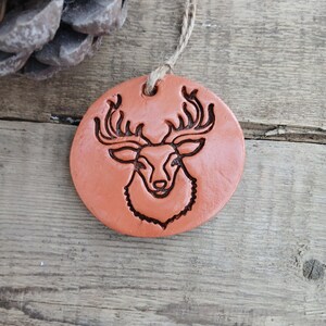 Puede incluir: Adorno redondo de color terracota con un grabado de la cabeza de un ciervo. El ciervo tiene grandes cuernos y una cara detallada. Una anilla de cuerda natural está unida para colgarlo. El adorno está sobre un fondo de madera rústica.