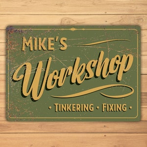 May include: A green metal sign with a distressed finish. The sign reads "Mike's Workshop" in yellow cursive lettering with the words "Tinkering" and "Fixing" in smaller yellow lettering below.