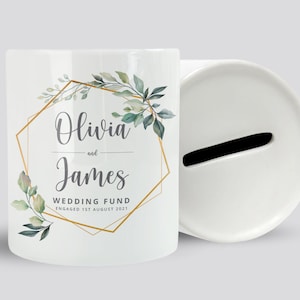 May include: White ceramic money box with a gold geometric frame and green foliage design. The text "Olivia and James Wedding Fund Engaged 1st August 2021" is printed on the front.
