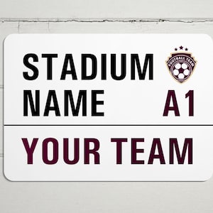 May include: White sign with black text that reads "Stadium Name A1 Your Team". A burgundy and gold soccer ball logo is in the top right corner.