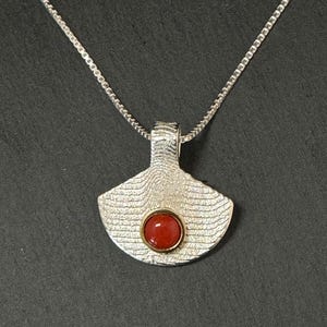 Può includere: Una collana d'argento con un ciondolo testurizzato con una pietra preziosa rossa. Il ciondolo ha la forma di un ventaglio e presenta una superficie testurizzata.