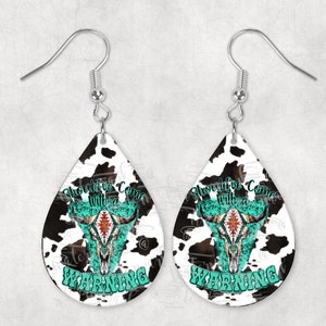 Puede incluir: Pendientes de lágrima con estampado de piel de vaca y diseño de calavera de toro turquesa y roja. Los pendientes tienen el texto "Should've Come With a Warning" en turquesa. Cuelgan de ganchos plateados.