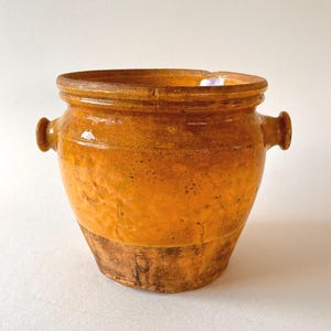 May include: A rustic, antique-style ceramic pot with a wide, open top and two small handles. The pot has a warm, yellow-orange glaze with darker brown tones at the base. The aged appearance suggests it is vintage or handcrafted.