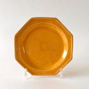 Può includere: Un piatto ottagonale in ceramica giallo senape. Il piatto ha una superficie strutturata e un bordo perlato. È esposto su un supporto in acrilico trasparente su sfondo bianco. Questo piatto è adatto per servire cibo.
