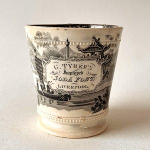 May include: A cream-colored ceramic cup featuring a black transfer-printed landscape design. The central cartouche displays the text G. TYRER'S Improved SODA FONT LIVERPOOL. The cup has a beaded rim and base, with a scene depicting a pagoda and a boat.