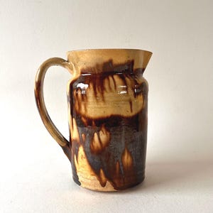 Op de afbeelding: Handgemaakte keramische kan met een gebogen handvat. De kan heeft een beige bovenkant met donkerbruin druipglazuur over een beige basis. Het aardewerk heeft een rustieke, ambachtelijke uitstraling, geschikt voor het serveren van dranken.