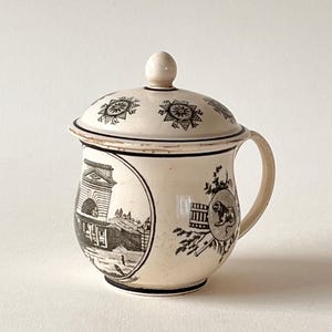 Pode incluir: Uma caneca de cerâmica cor de creme com tampa, decorada com ilustrações impressas em preto. O corpo apresenta uma cena circular de um edifício clássico e um emblema de leão. A tampa tem motivos em forma de estrela e um pequeno puxador.