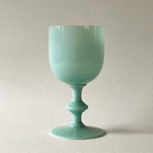 May include: A smooth, opaque light mint green glass goblet featuring a rounded bowl, a slender stem with a central decorative knob, and a wide, circular base. The surface of the glass has a soft, glossy finish that reflects light evenly.