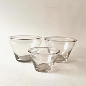 Pode incluir: Três tigelas de vidro transparente de tamanhos variados. As tigelas têm uma forma alargada com uma borda arredondada. A tigela menor está em primeiro plano, com duas tigelas maiores atrás dela. As tigelas estão vazias e sobre uma superfície branca.