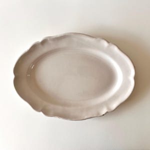 Może przedstawiać: Owalny półmisek ceramiczny z gładkim, kremowobiałym szkliwem. Naczynie posiada falisty, dekoracyjnie wykończony brzeg na całym obwodzie, co nadaje tej funkcjonalnej części zastawy stołowej miękki i zaokrąglony kształt.